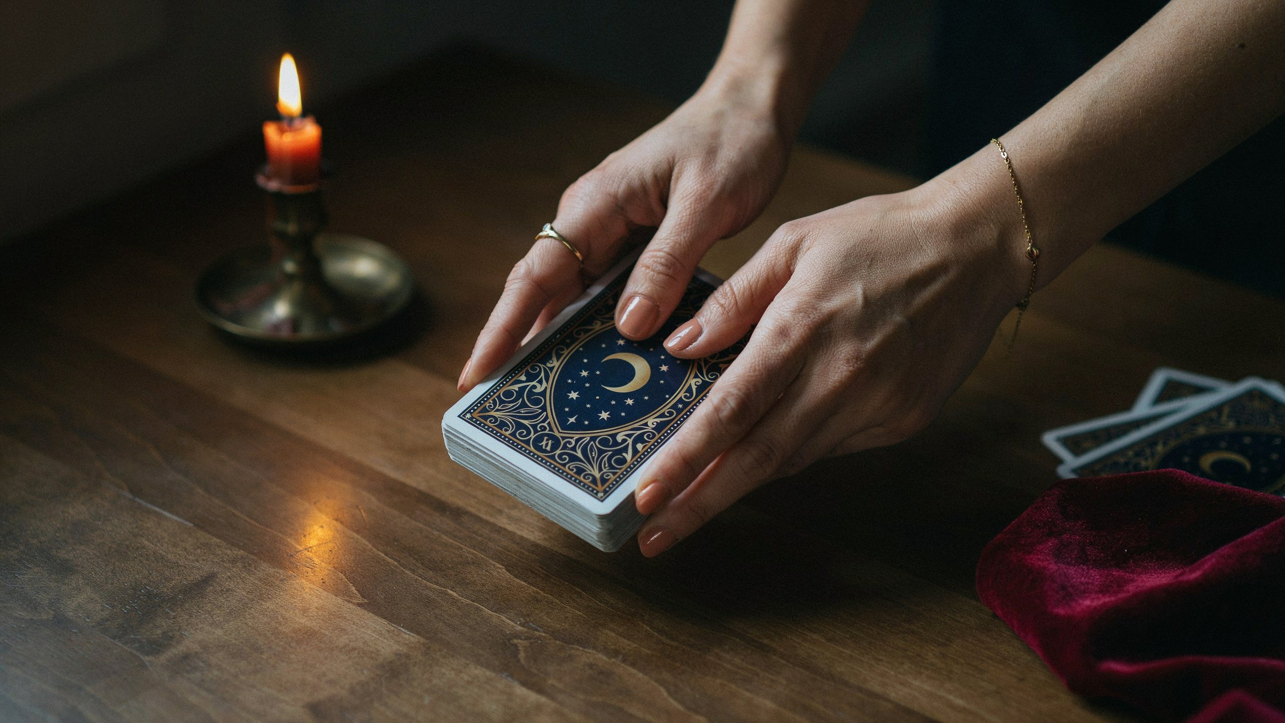 Maos embaralhando um baralho de tarot com carta de fundo azul e lua dourada vela acesa ao lado sobre mesa de madeira